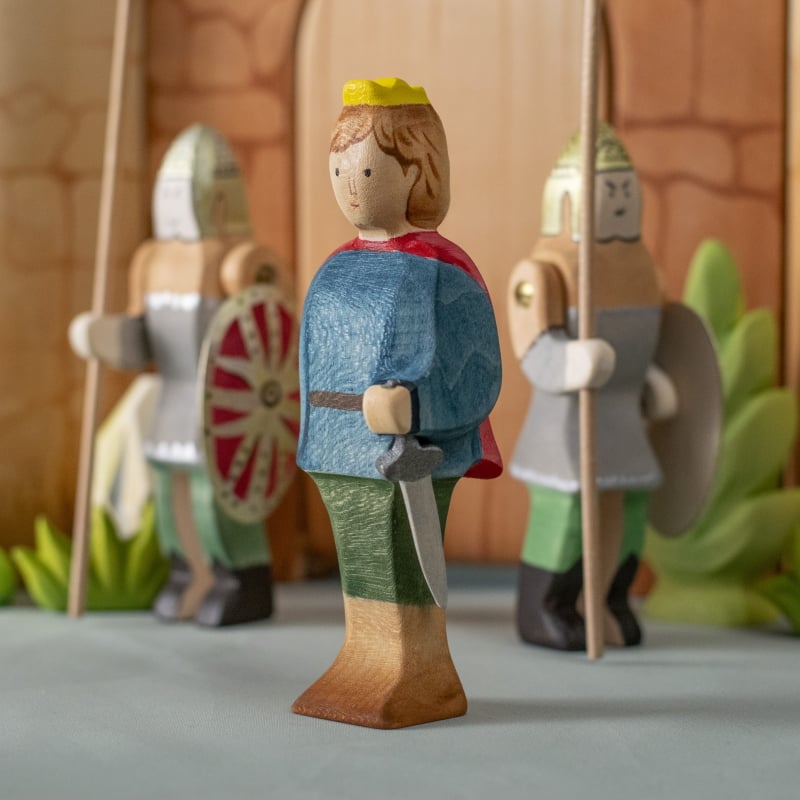 The Bumbu Handmade Wooden Prince, in a blue shirt, green pants, red cape, and yellow crown, stands before two armored guards with a medieval castle toy backdrop—an ideal set to inspire imaginative play.