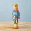 The Bumbu Handmade Wooden Prince, featuring a yellow crown, red cape, blue top, green pants, and sword, stands on a light wooden surface with a plain light blue background.