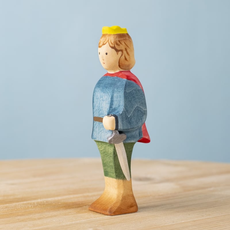 The Bumbu Handmade Wooden Prince, featuring a yellow crown, red cape, blue top, green pants, and sword, stands on a light wooden surface with a plain light blue background.