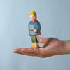 A hand with a gold ring holds the Bumbu Handmade Wooden Prince, a handcrafted figure wearing a yellow crown, blue shirt, green pants, and red cape, set against a light blue background.