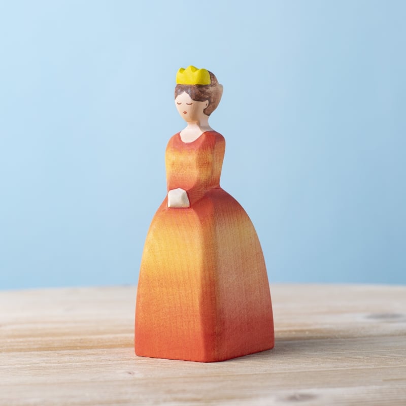 The Bumbu Handmade Wooden Princess is a small, sustainable wooden figure of a woman in an orange-red gradient dress with a yellow crown. Ideal for Waldorf-inspired play and eco-friendly toy collections.
