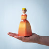 The Bumbu Handmade Wooden Princess, featuring a woman in a yellow-orange dress and crown, is balanced on an open palm against a plain light blue background.