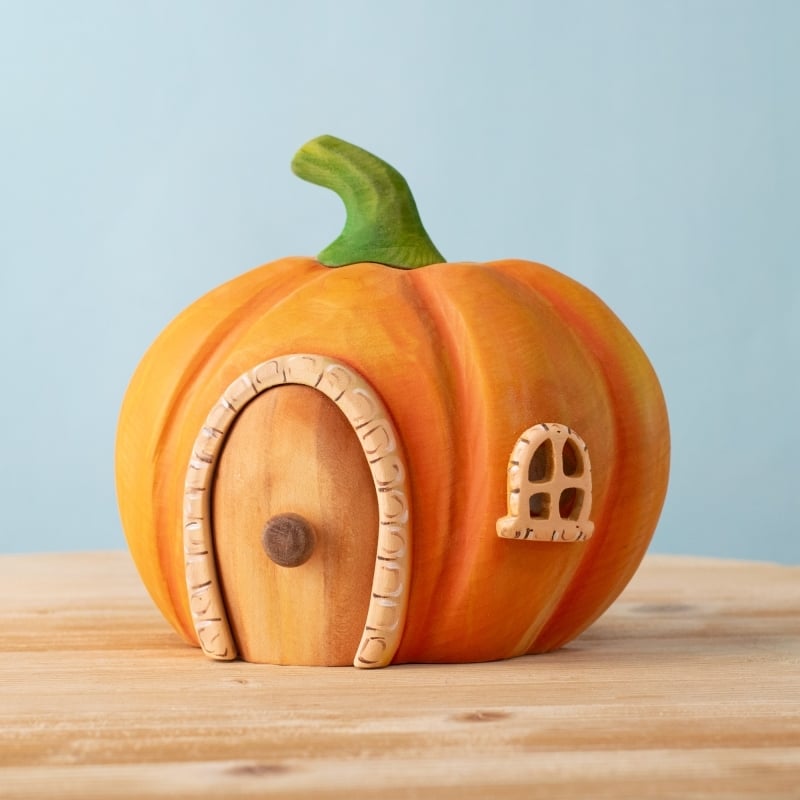 The Bumbu Handmade Wooden Pumpkin House, featuring a green stem, wooden door with round handle, and Montessori-inspired play details, sits on a wooden surface against a light blue background.