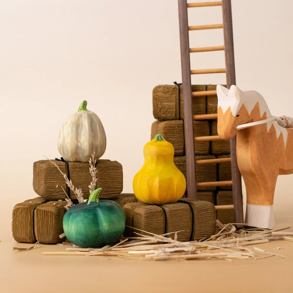 Three wooden pumpkins in green, yellow, and white sit on brown blocks by a toy horse and ladder—a charming Bumbu Handmade Wooden Pumpkin Patch Set for Montessori play in a neutral setting.