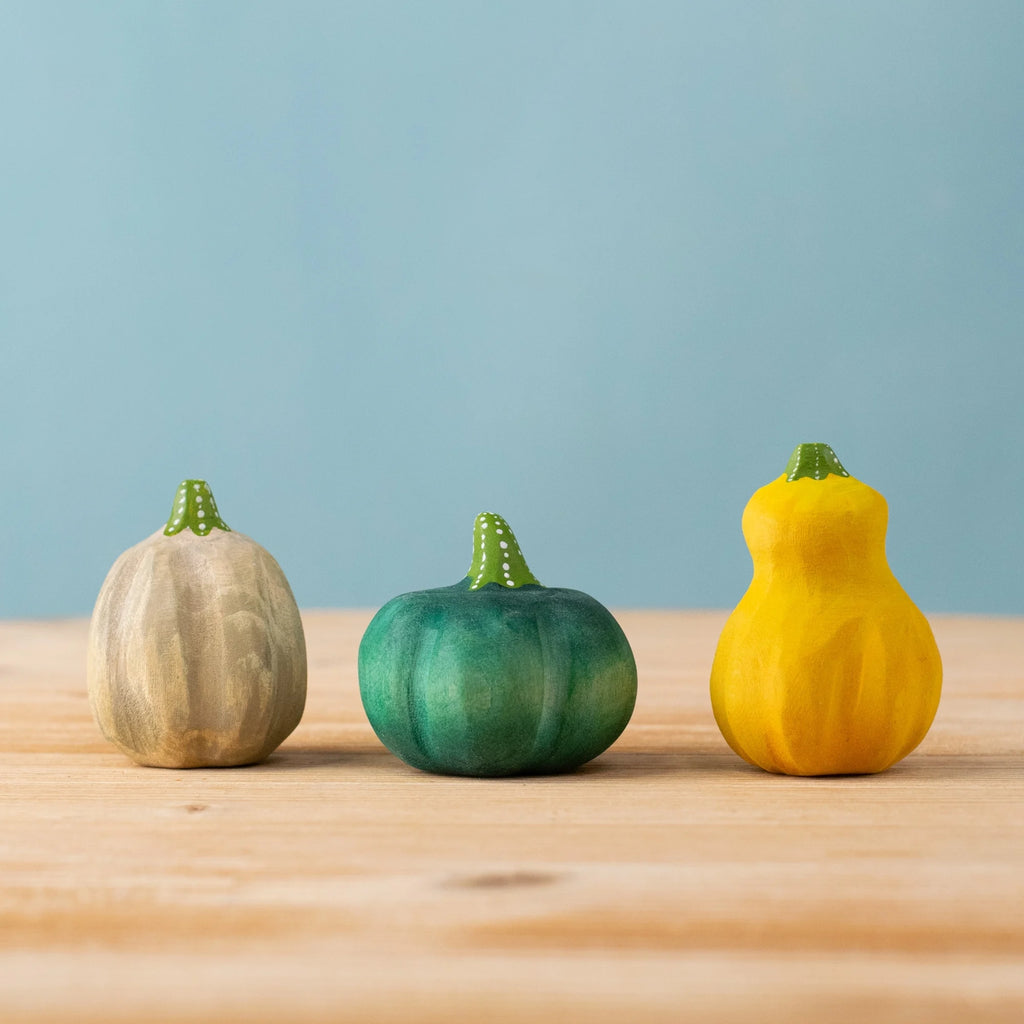 Three small Bumbu Handmade Wooden Pumpkin Patch Set pumpkins in gray, green, and yellow with green stems are arranged in a row—a charming Montessori play set on a wooden surface against a light blue background.