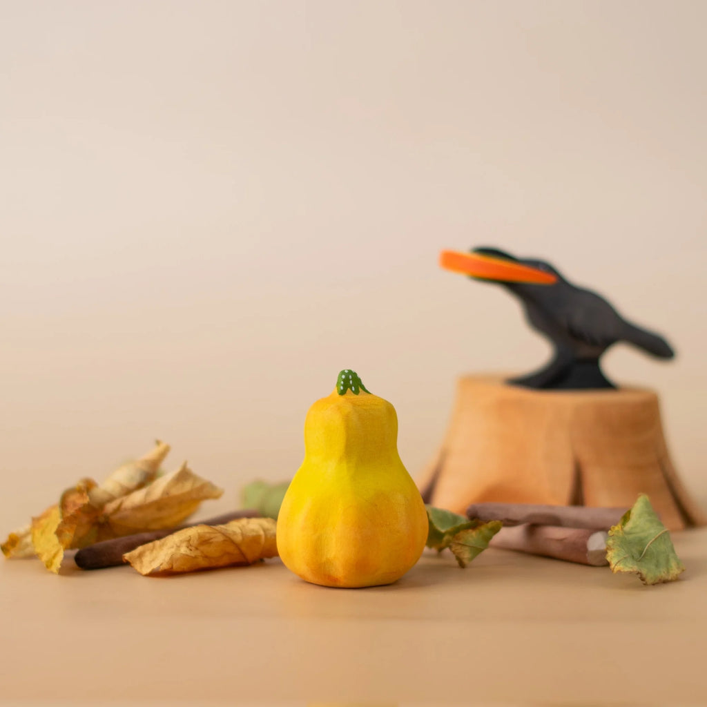 A small yellow gourd among dried leaves and twigs, with a black bird figurine in the background—perfect for Montessori play or paired with the Bumbu Handmade Wooden Pumpkin Patch Set.