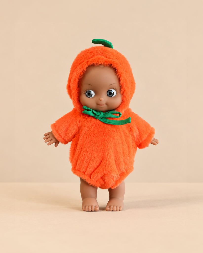 A Minikane Mini Pancha Doll - Maia Celebrating with Pumpkin Halloween Outfit wearing a fuzzy, hooded orange pumpkin costume with a green stem on top.