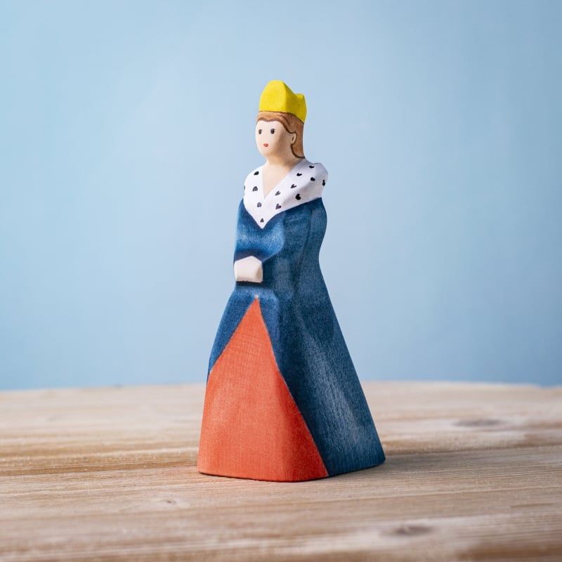 The Bumbu Handmade Wooden Queen, featuring a yellow crown, blue and red dress, and white fur-trimmed cape, stands on a wooden surface against a light blue background.