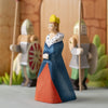 The Bumbu Handmade Wooden Queen, dressed in a blue and red gown with a yellow crown, stands before two armored toy knights with shields against a castle backdrop.