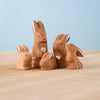 The Bumbu Handmade Wooden Brown Bunny Rabbit Family Set features six small, handcrafted wooden bunny figurines with white details and heart shapes, arranged on a light wood surface against a pale blue background—perfect for Montessori collections.