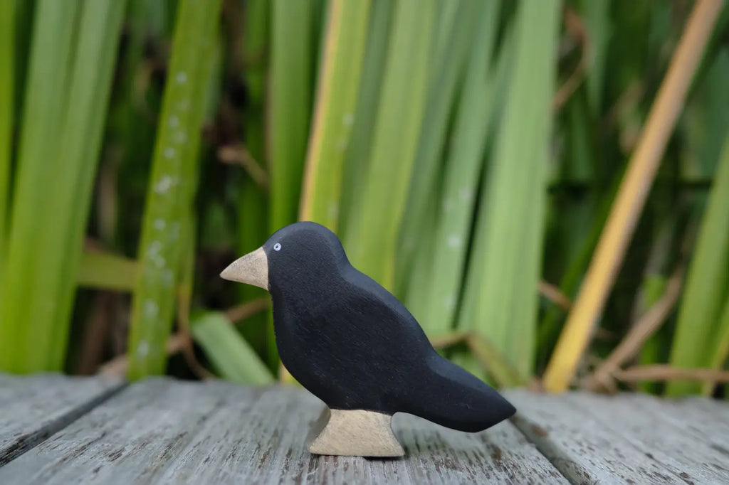 A handmade Holzwald Bird - Raven figurine with black wood, a light-colored beak and base, standing on a wooden surface against a background of tall green grass.