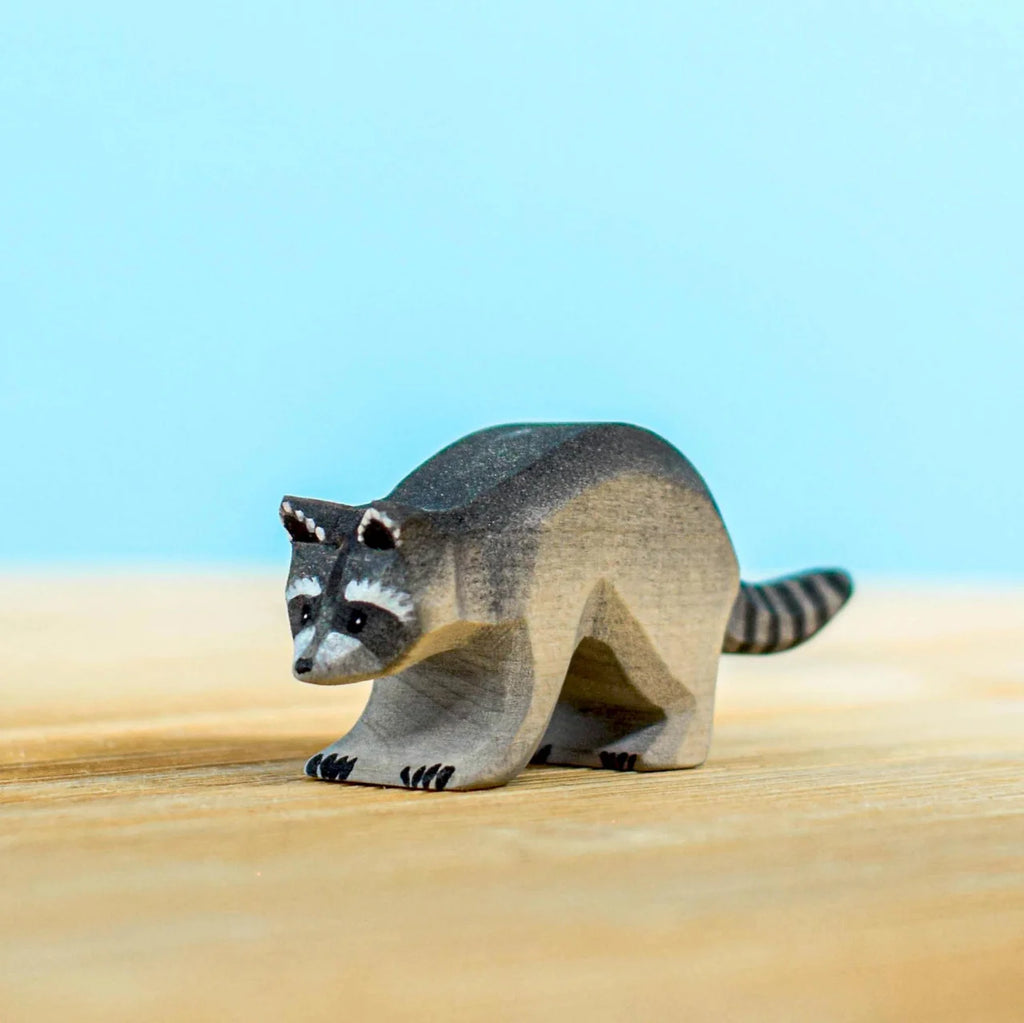 A figurine from the Bumbu Wooden Raccoon Set stands on a wooden surface against a light blue background. As part of an imaginative play handcrafted set, it features intricate details with black and white markings characteristic of a raccoon.