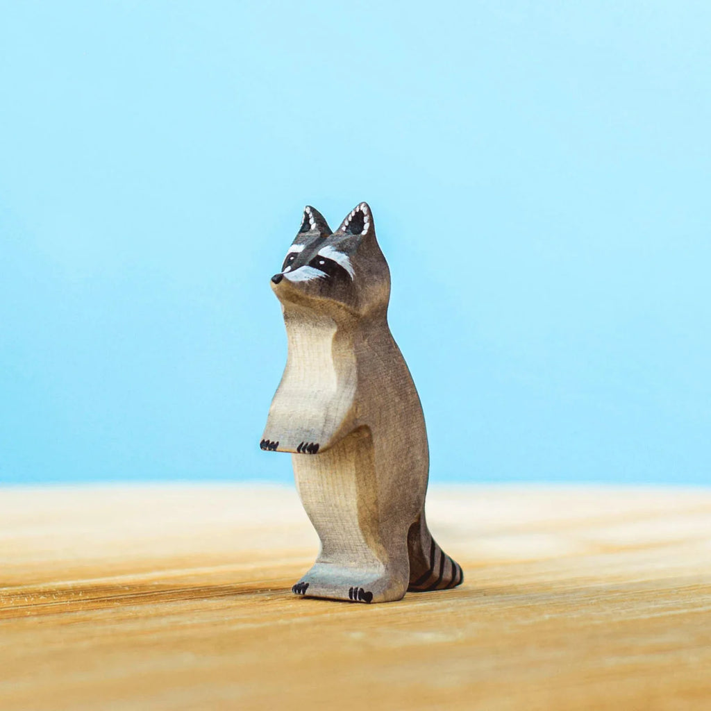 A small wooden raccoon figurine from the Bumbu Wooden Raccoon Set stands on a light wood surface against a light blue background. Handcrafted with black and gray details, it depicts the characteristic mask and ringed tail, perfect for imaginative play as part of your Wooden Raccoon Family collection.