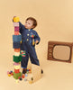 A toddler in a denim jumpsuit is stacking Raduga Grez Big Cube Block Set - Colorful, building a tall tower, with additional cubes scattered on the floor and a toy stove in the background.