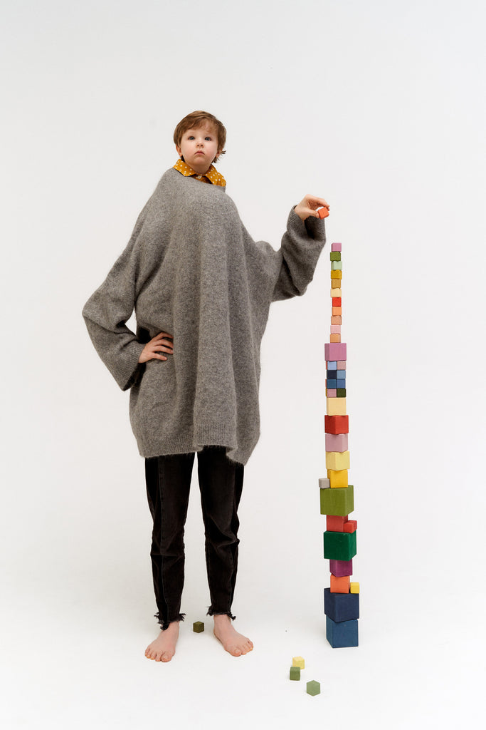 A young boy in an oversized grey sweater and black pants stands barefoot next to Raduga Grez | Big Cube Block Set - Colorful. He appears serious and is pointing at the top block.