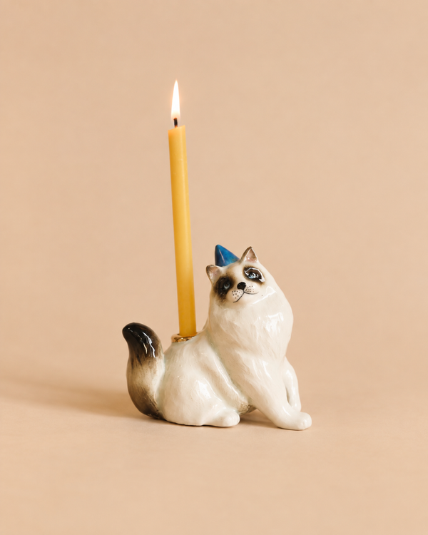 A whimsical Ragdoll Cat Cake Topper—featuring a ceramic cat figurine wearing a small blue party hat and holding a lit yellow candle—is set against a beige background, making it the perfect cat-themed decoration for your next celebration.