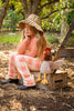 A young girl with long hair sits on a wooden crate outdoors, wearing a wide-brimmed straw hat, an orange long-sleeve shirt, checkered pants, and black boots. She rests her chin on her hand, looking pensive. Beside her is Randy the Rooster Stuffed Animal. Trees and foliage are in the background.