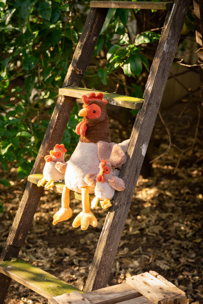 Randy the Rooster Stuffed Animal, complete with two attached chicks, sits on a wooden ladder outdoors. The ladder is set against a backdrop of lush green foliage, illuminated by warm sunlight to create a cozy and inviting scene.