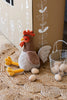 Randy the Rooster Stuffed Animal, with its soft brown and beige body, orange comb, and yellow feet, sits cozily on a woven rug. Next to it, a white wire basket holds beige eggs. The backdrop showcases a cardboard structure adorned with white flower illustrations.