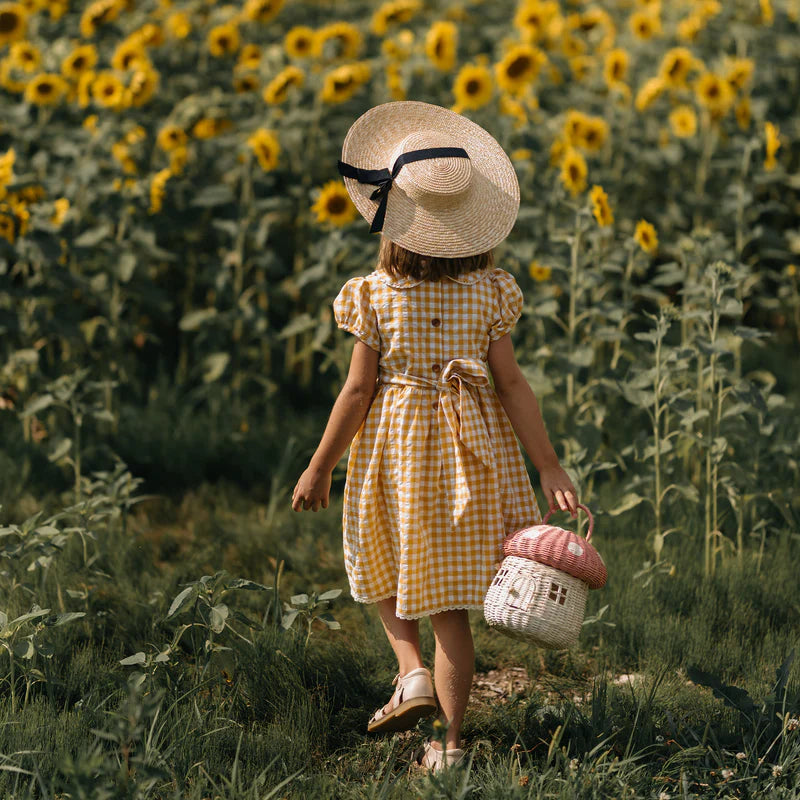 A young girl in a checkered dress and straw hat, carrying an Olli Ella Mushroom Basket in Pink, walks through a sunflower field. Her back is to the camera, highlighting a sense of exploration.