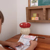 A child colors at a wooden desk with the Olli Ella Mushroom Basket With Coloring Pencils- Red. Twig pencils and the woven basket are on the desk, with wooden alphabet blocks and a small shelf above.
