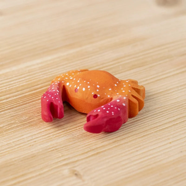 The Bumbu Handmade Wooden Red Crab, a small handcrafted figurine with red claws and white spots, sits on a light wooden surface.