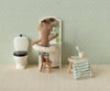 A stuffed animal wearing a swim ring stands on a stool in front of a vanity mirror in a dollhouse bathroom with a toilet, striped towel, the Maileg Toothbrush Set in Bag, Mouse - Blue Stripe, and other charming dollhouse accessories.