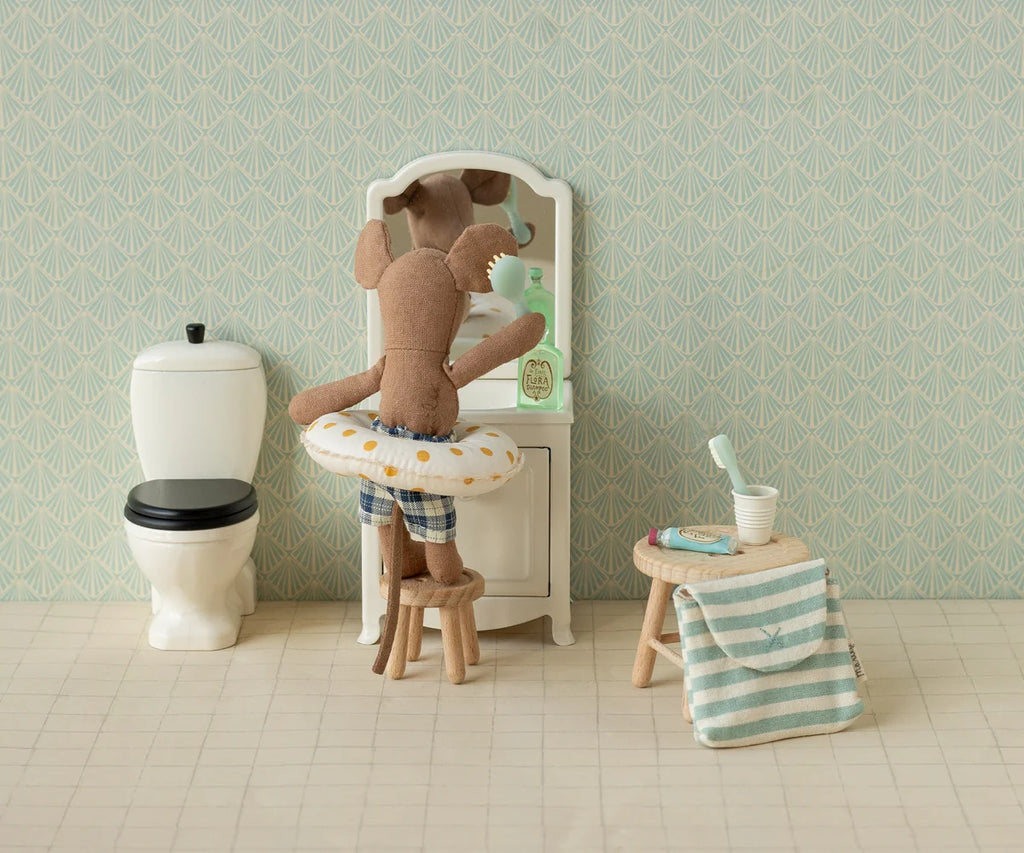A stuffed animal wearing a swim ring stands on a stool in front of a vanity mirror in a dollhouse bathroom with a toilet, striped towel, the Maileg Toothbrush Set in Bag, Mouse - Blue Stripe, and other charming dollhouse accessories.