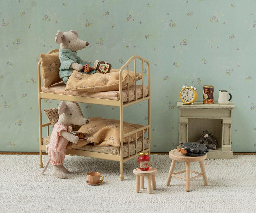 Tucked in the whimsical Maileg Bunk Bed, Mouse - Wheat (ships in 1-2 weeks), two pajama-clad mice enjoy a cozy sleepover—one reading atop the upper bunk while the other sits up below. Styled like a charming Mouse hole Farmhouse, this imaginative set features a tiny nightstand with food, a clock, and stove, along with miniature stools and cups arranged on a patterned rug—perfect for creative play.