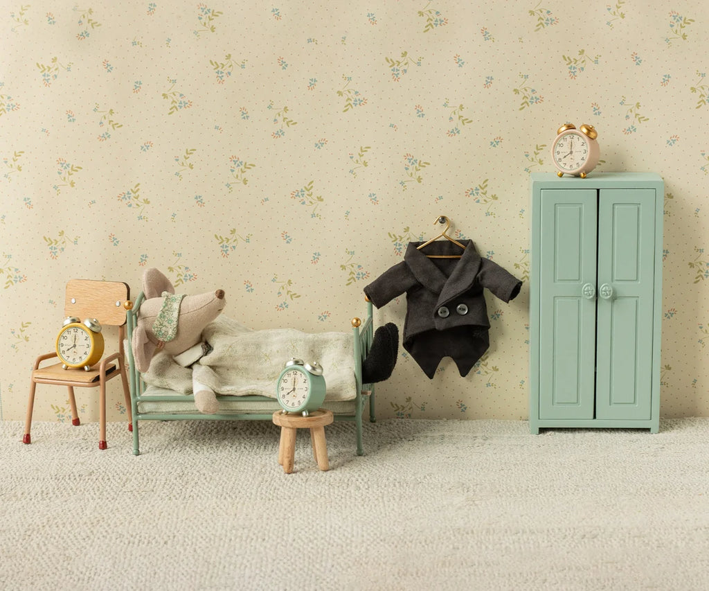 A collectible Maileg Wedding Mice Couple in Box, Mum and Dad, lies in a bed beside a wardrobe, hanging coat, chair, and stool. Four alarm clocks are placed on the furniture within this miniature, vintage-style bedroom—perfect for collectors of Maileg wedding mice or as a charming wedding gift.