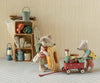 Two toy mice, dressed for an outing, pack camping gear. One pulls a Maileg Wagon, Mouse – Red (estimated arrival mid September), filled with snacks and thermoses, while the other holds a striped floatie. Camping supplies and cookware—along with other charming mouse family accessories—are neatly stacked on a wooden shelf nearby.
