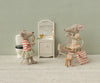 Two toy mice in a miniature bathroom share a bedtime routine: one stands with a magnifying glass near the sink and toilet, while the other sits on a chair in a floral dress. Among their charming accessories are a mirror, toothbrush, and striped bag from the Maileg Toothbrush Set in Bag, Mouse – Coral Stripe.