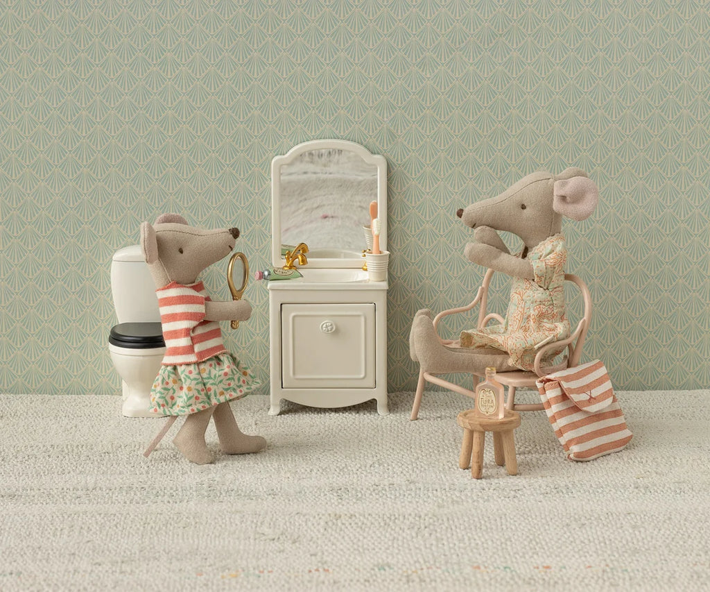 Two toy mice in a miniature bathroom share a bedtime routine: one stands with a magnifying glass near the sink and toilet, while the other sits on a chair in a floral dress. Among their charming accessories are a mirror, toothbrush, and striped bag from the Maileg Toothbrush Set in Bag, Mouse – Coral Stripe.