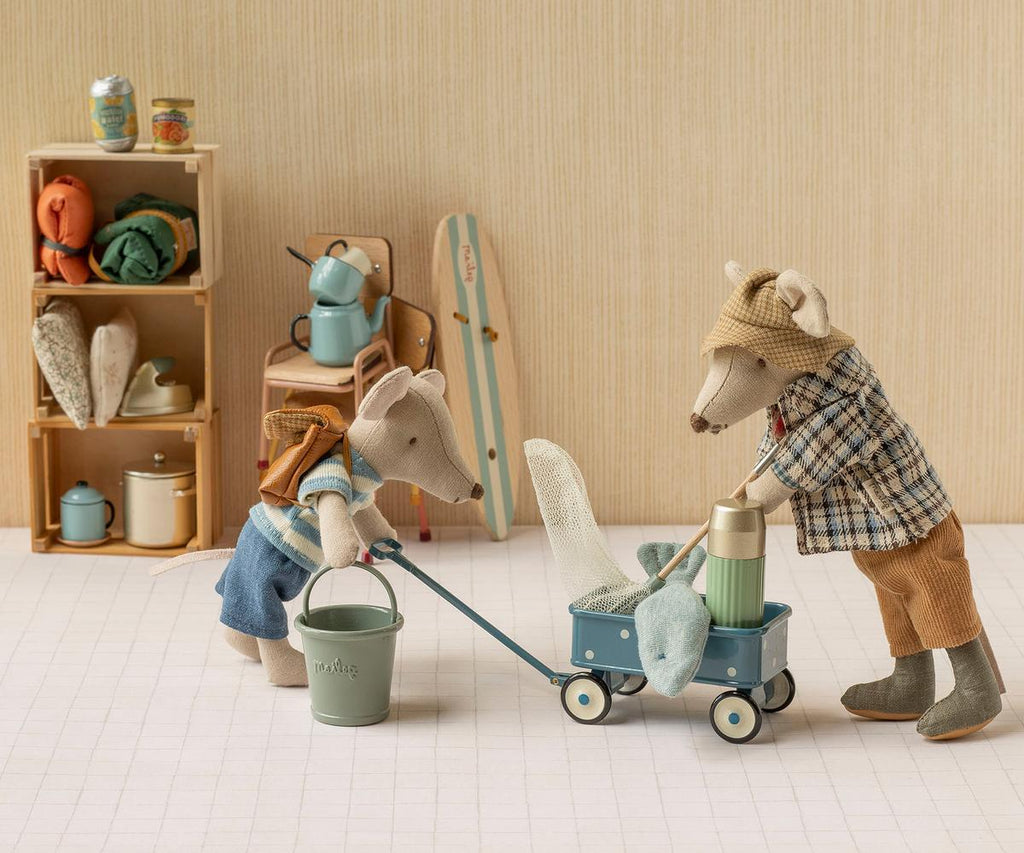 Two toy mice in vintage-style clothes play with the PRE-ORDER: Maileg Wagon, Mouse – Blue (estimated arrival mid September) and a toy bucket. Behind them, a shelf displays miniature household items, mouse family accessories, and a skateboard. The scene is set against a beige background.