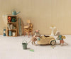 Two toy mice in clothes play with the PRE-ORDER: Maileg Car, Mouse - Wheat (Estimated Arrival Mid August), a small metal car. Behind them are shelves, stacked items, a surfboard, chairs, a small sink, and tiny household objects—all set against a beige wall and white checked floor.