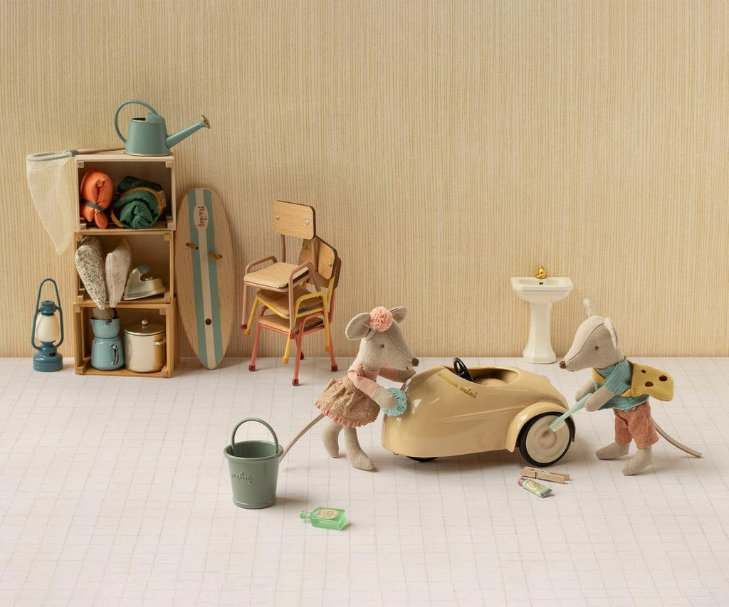 Two toy mice in clothes play with the PRE-ORDER: Maileg Car, Mouse - Wheat (Estimated Arrival Mid August), a small metal car. Behind them are shelves, stacked items, a surfboard, chairs, a small sink, and tiny household objects—all set against a beige wall and white checked floor.