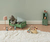 A cozy children's room scene with a teddy bear in a green bed, toy mice riding on the PRE-ORDER: Maileg Car, Mouse - Wheat (Estimated Arrival Mid August), a fluffy white rug, a lamp, and a small green shelf against a pale wall.