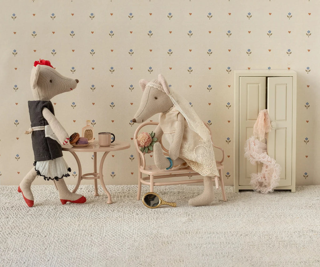 The Maileg Wedding Mice Couple in Box, Mum and Dad, are adorably dressed as bride and groom, seated by a table with treats. A nearby wardrobe adorned with a pink scarf and a handheld mirror on the floor complete the scene, set against patterned wallpaper. This enchanting collectible makes a thoughtful wedding gift for any Maileg fan.