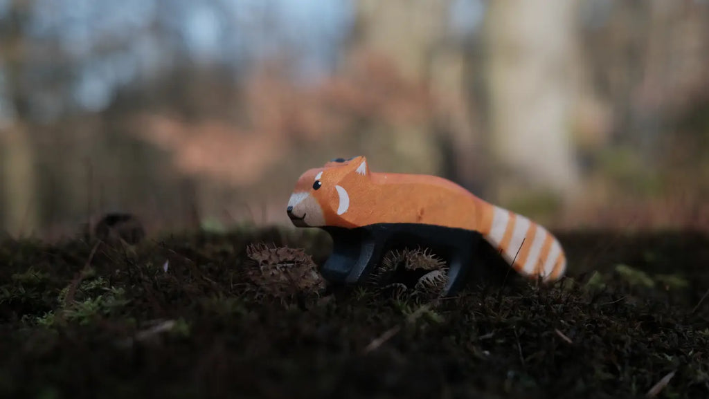 The Handmade Holzwald Red Panda, a wooden figurine with orange and white markings, stands on mossy ground outdoors with a blurred forest background—a charming example of Holzwald's craftsmanship.