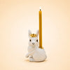 Introducing the Royal White Rabbit Cake Topper – Final Sale: a handcrafted collectible featuring a white rabbit figurine adorned with a gold crown and carrying a tall, glowing yellow candle on its back, beautifully set against a simple beige backdrop.