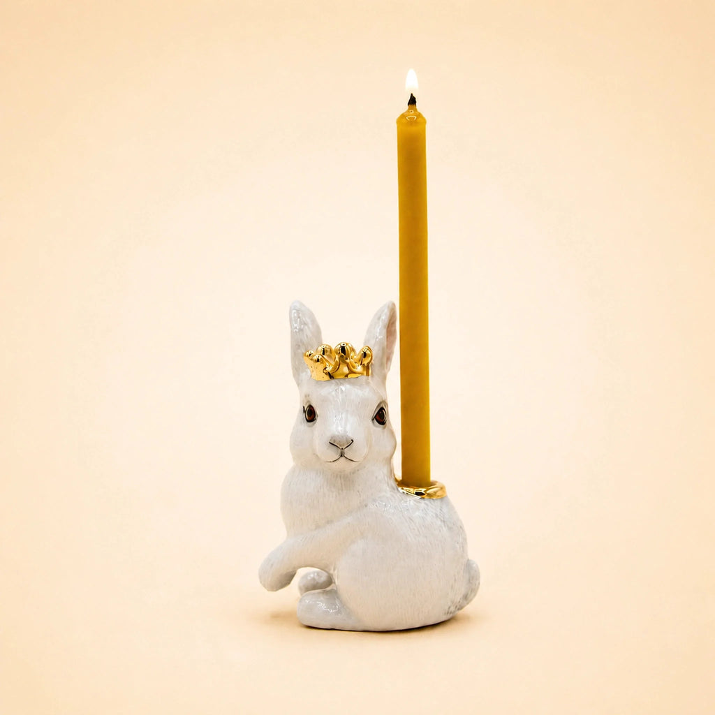 Introducing the Royal White Rabbit Cake Topper – Final Sale: a handcrafted collectible featuring a white rabbit figurine adorned with a gold crown and carrying a tall, glowing yellow candle on its back, beautifully set against a simple beige backdrop.