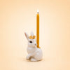 A charming handcrafted collectible, the Royal White Rabbit Cake Topper features a white rabbit figurine adorned with a gold crown, holding a lit yellow candle on its back against a soft beige background.