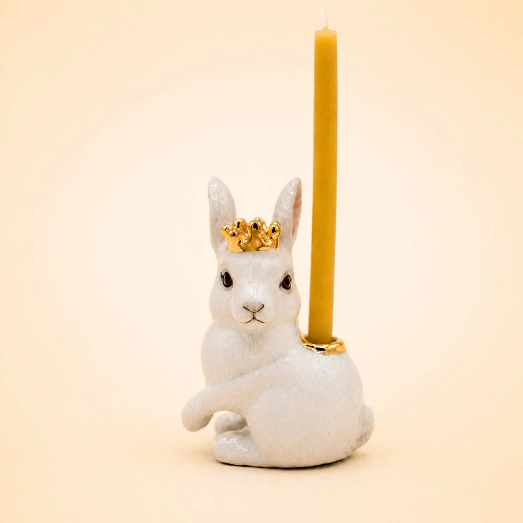 A Royal White Rabbit Cake Topper, featuring a handcrafted white ceramic rabbit adorned with a gold crown and carrying a yellow candle on its back, makes a charming accent for any space. Shown here against a plain light beige background.