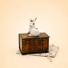 The “Royal White Rabbit Cake Topper – Final Sale,” a petite white rabbit figurine adorned with a gold crown, sits elegantly atop a vintage-style keepsake treasure chest, which is displayed on a beige drawstring pouch decorated with star motifs, all set against a plain cream background.