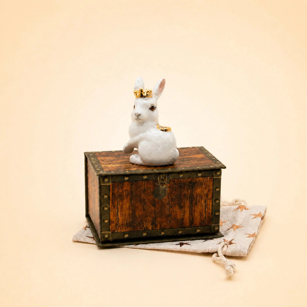 The “Royal White Rabbit Cake Topper – Final Sale,” a petite white rabbit figurine adorned with a gold crown, sits elegantly atop a vintage-style keepsake treasure chest, which is displayed on a beige drawstring pouch decorated with star motifs, all set against a plain cream background.