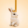 The Royal White Rabbit Cake Topper - Final Sale features a white ceramic rabbit figurine adorned with a gold crown and holding a slender, upright yellow stick. Set against a soft, light beige background, this elegant porcelain cake topper adds a refined touch to any keepsake chest or display collection.