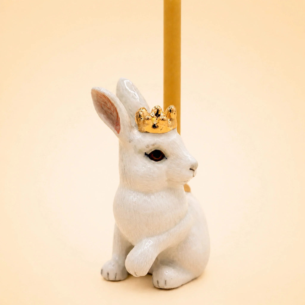 The Royal White Rabbit Cake Topper - Final Sale features a white ceramic rabbit figurine adorned with a gold crown and holding a slender, upright yellow stick. Set against a soft, light beige background, this elegant porcelain cake topper adds a refined touch to any keepsake chest or display collection.
