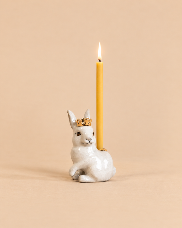 Introducing the “Royal White Rabbit Cake Topper - Final Sale”: a handcrafted collectible ceramic bunny figurine adorned with a gold crown, carrying a lit yellow candle on its back, beautifully presented against a plain beige background.