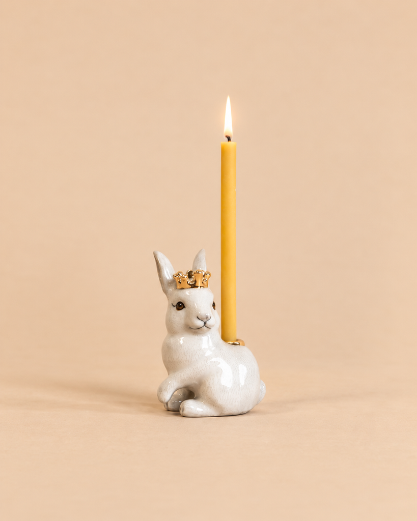 Introducing the “Royal White Rabbit Cake Topper - Final Sale”: a handcrafted collectible ceramic bunny figurine adorned with a gold crown, carrying a lit yellow candle on its back, beautifully presented against a plain beige background.