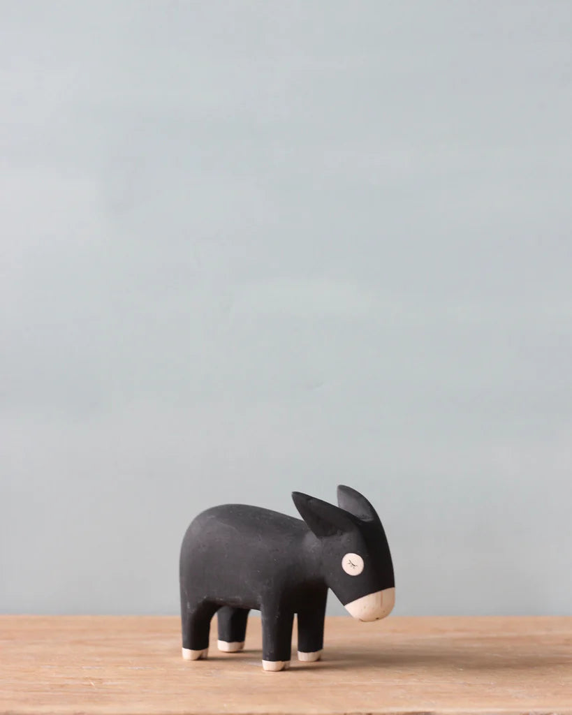A small black Handmade Tiny Wooden Farm Animals - Donkey figurine, with white details on its face and ears, stands on a wooden surface against a plain light blue background.